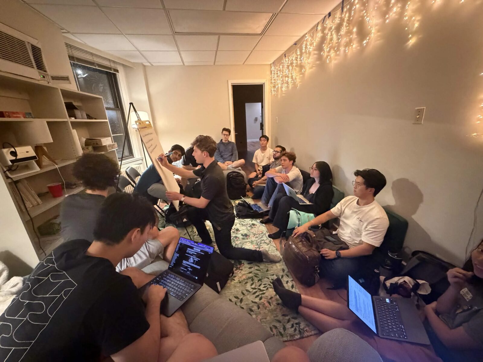 Kneeling at the flip chart, marker in hand, with the group watching from the couches and the floor
