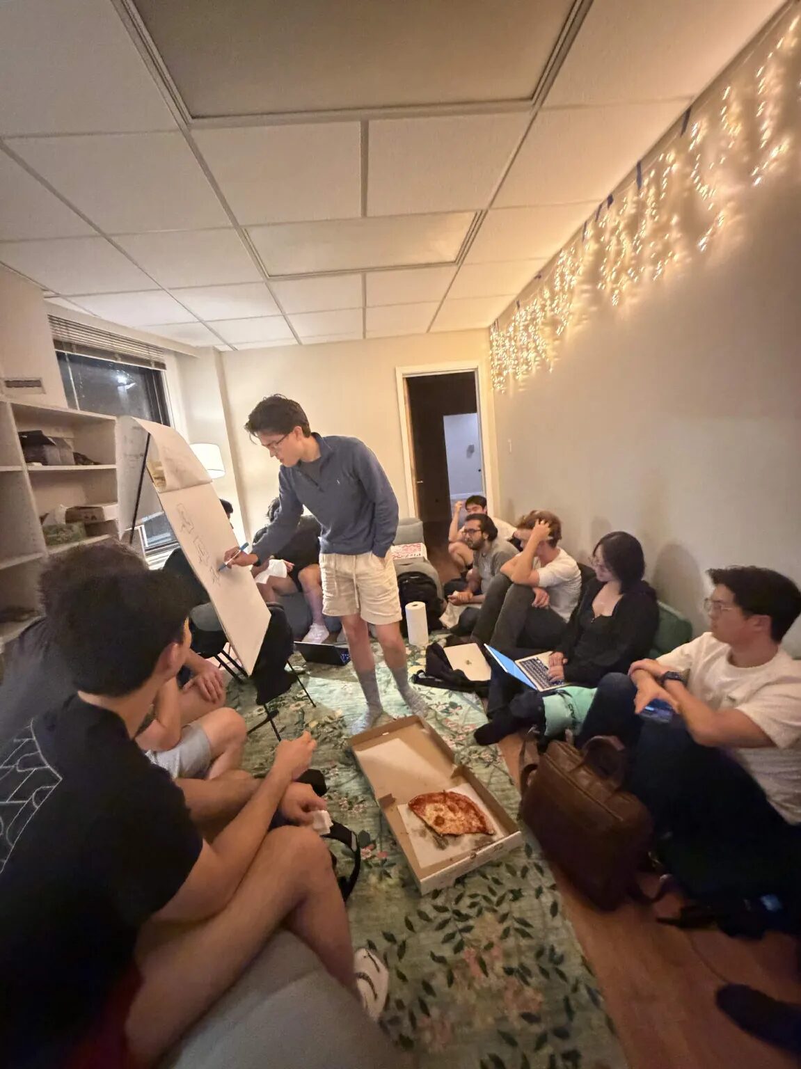 Standing at the flip chart, sketching, with a pizza box open on the rug